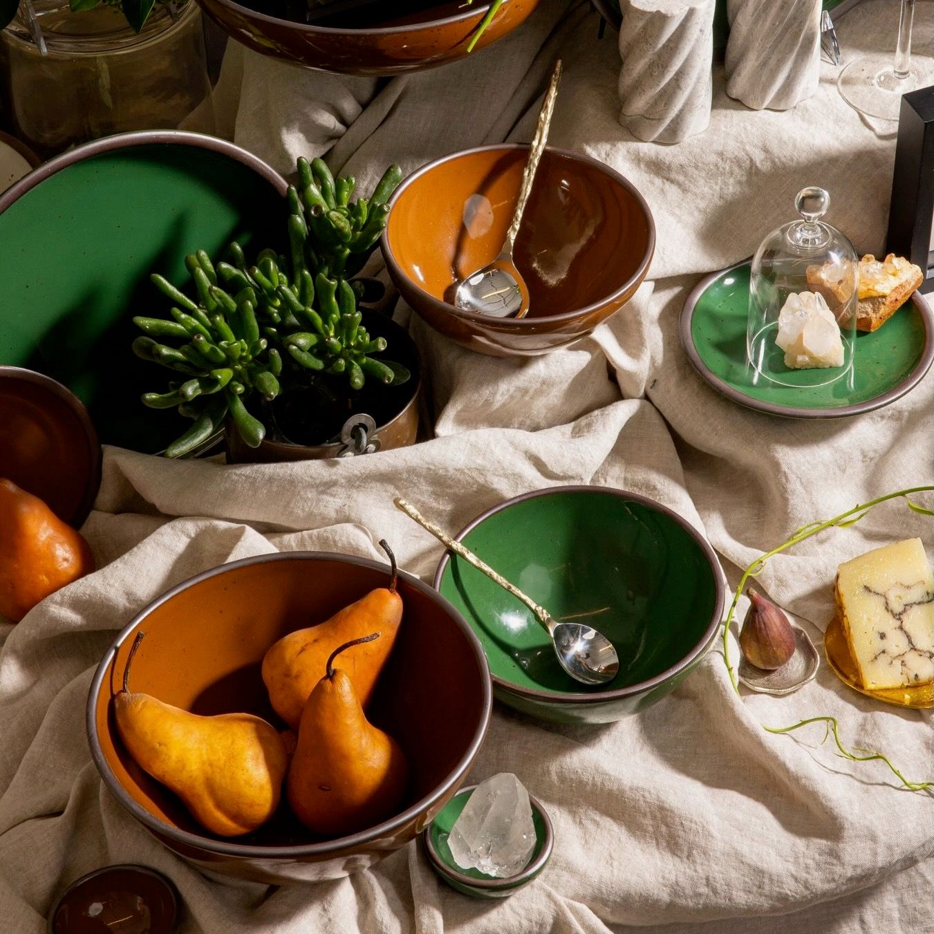 An artful arrangement of ceramic bowls in glossy green and brown colors on a table with a large tablecloth, plant, pears and more.