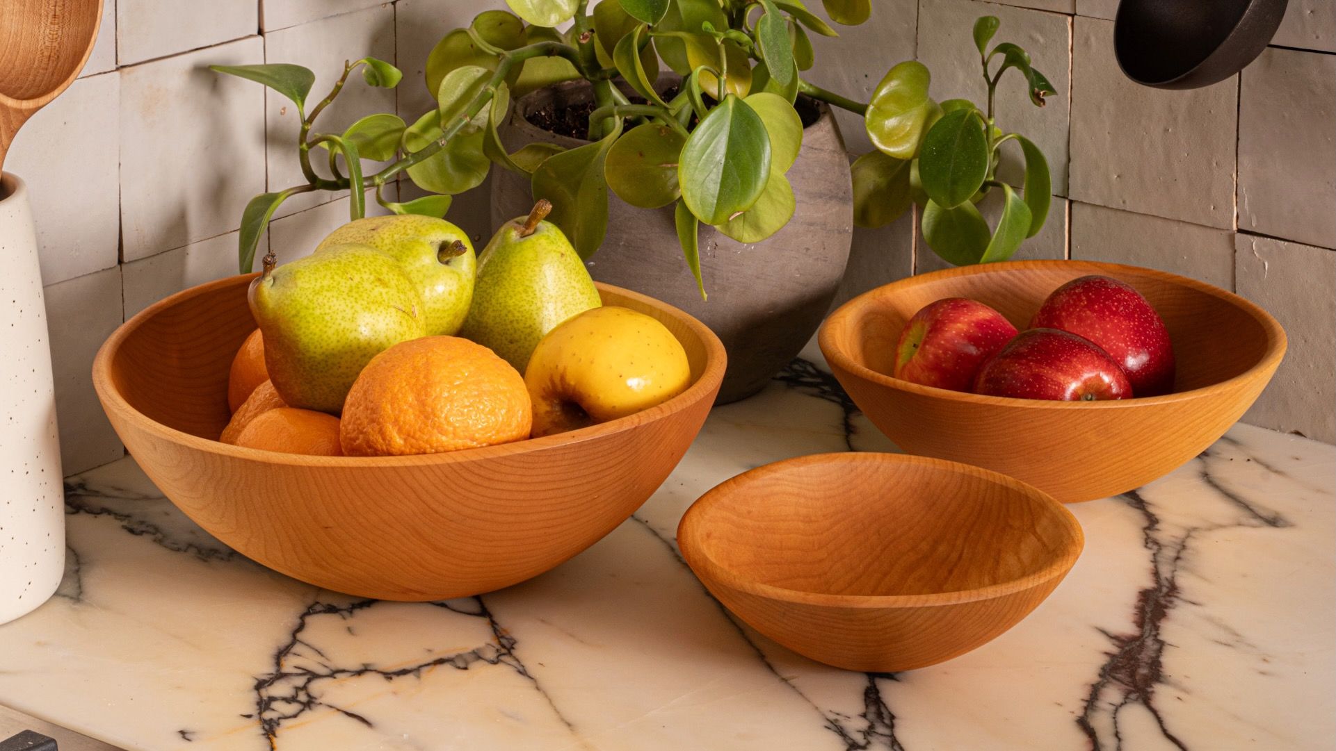 Wooden bowls filled with pears, oranges, and apples on a marble countertop beside a potted plant in a tiled kitchen.