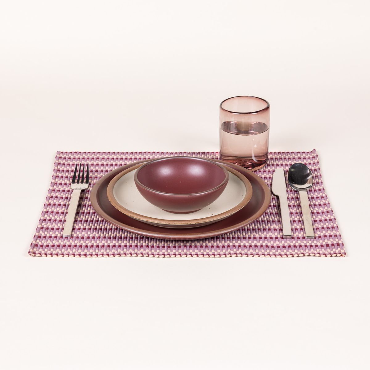 A place setting featuring a striped woven placemat with plum and blush colors, plates to match, a glass, and silverware.