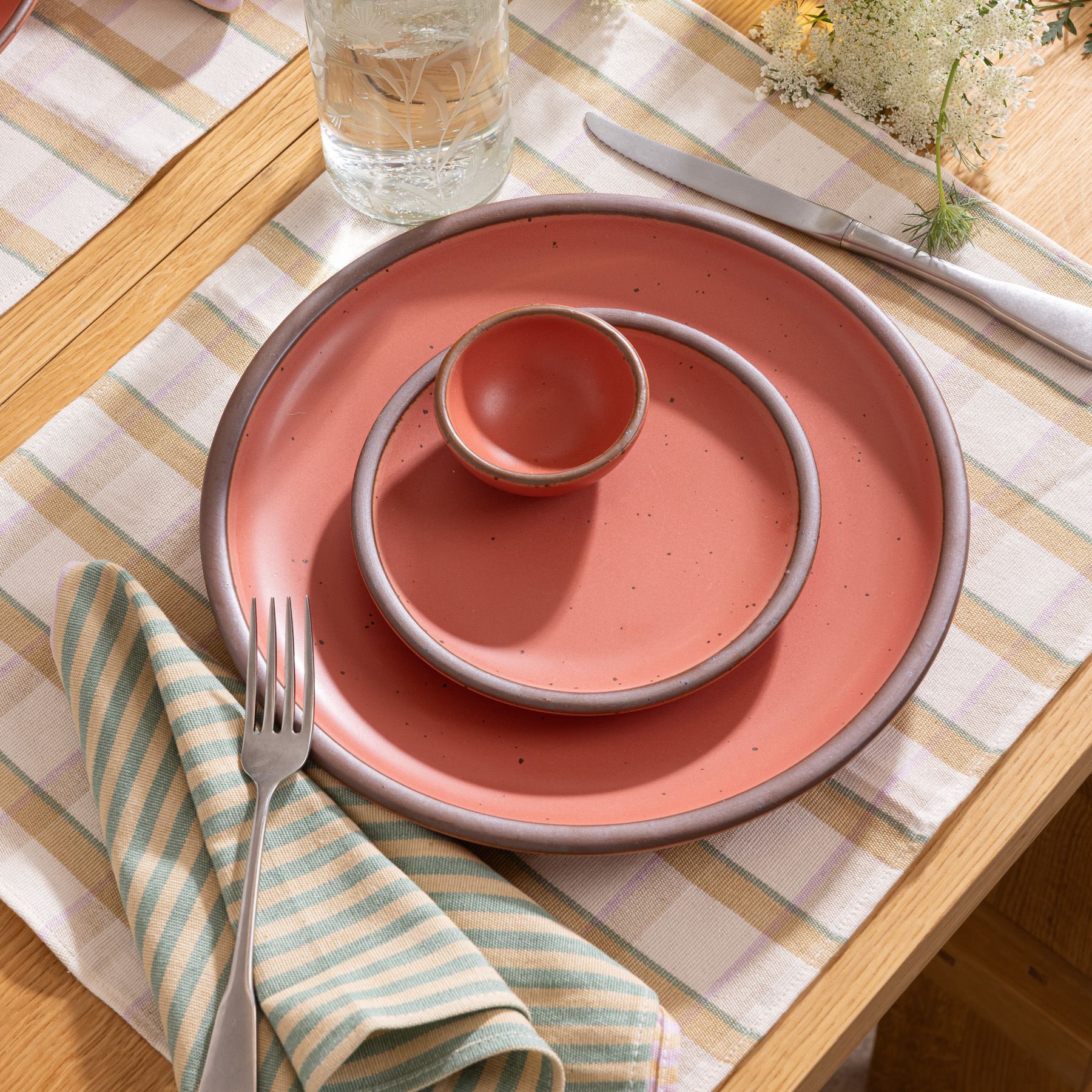 A place setting with watermelon pink ceramic dinner plate, side plate, and tiny bowl, against a pastel and neutral napkin and placemat.