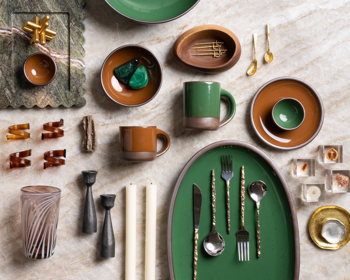 Flat lay of glossy green and brown ceramic dishes, mugs, utensils, glassware, candles, and decorative objects on marble.