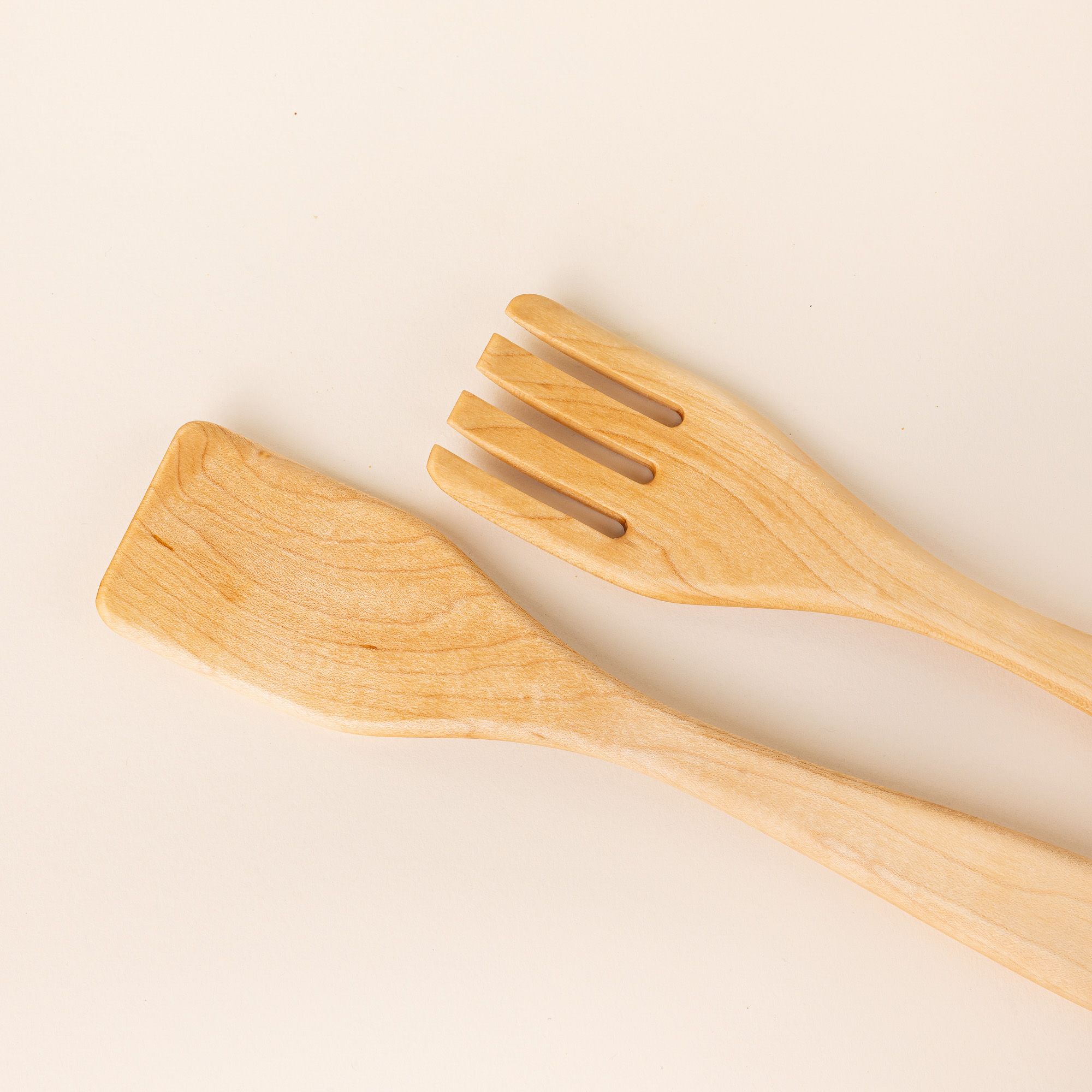 Closer look at the end of two long maple wood serving utensils. The left utensil has a shape like a spatula, the right has a shape like a fork.