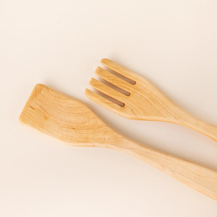 Closer look at the end of two long maple wood serving utensils. The left utensil has a shape like a spatula, the right has a shape like a fork.