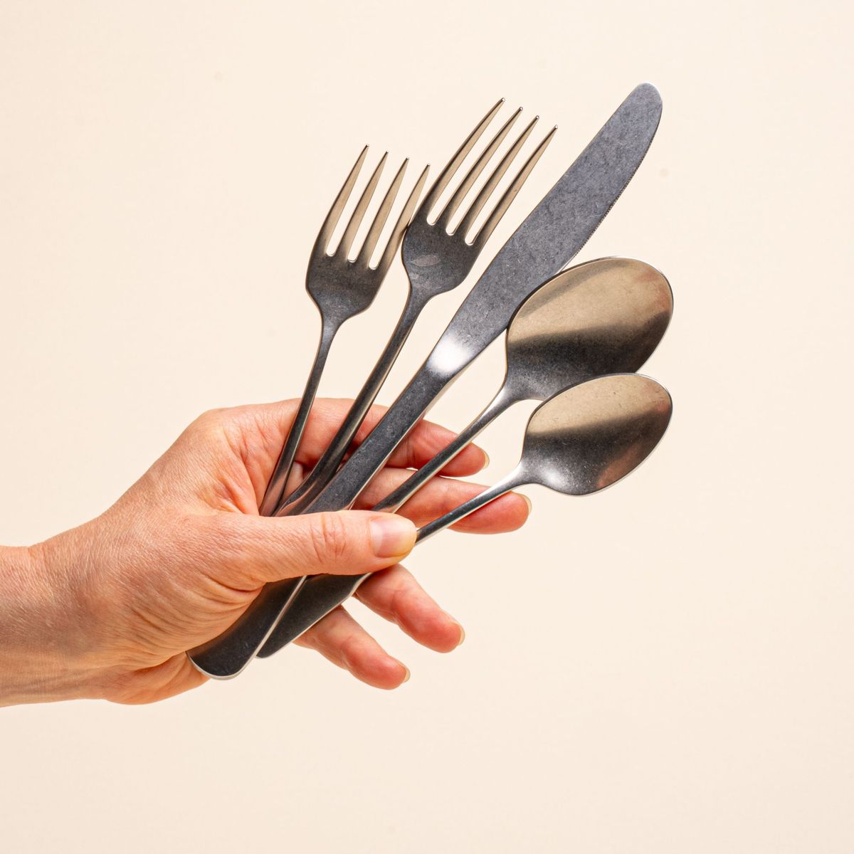 A hand holds a stainless steel five-piece flatware including 2 forks, 2 spoons, and 1 knife, in a tumbled matte finish and classic design