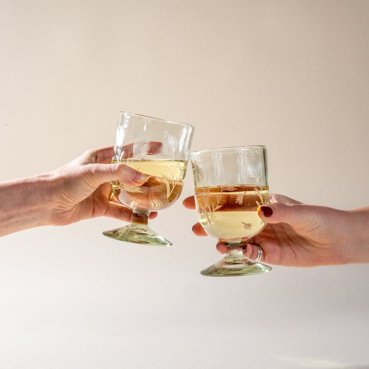 Two hands each hold a clear short wine glass made of recycled glass with etched thistle designs on it, filled with white wine, and cheers.