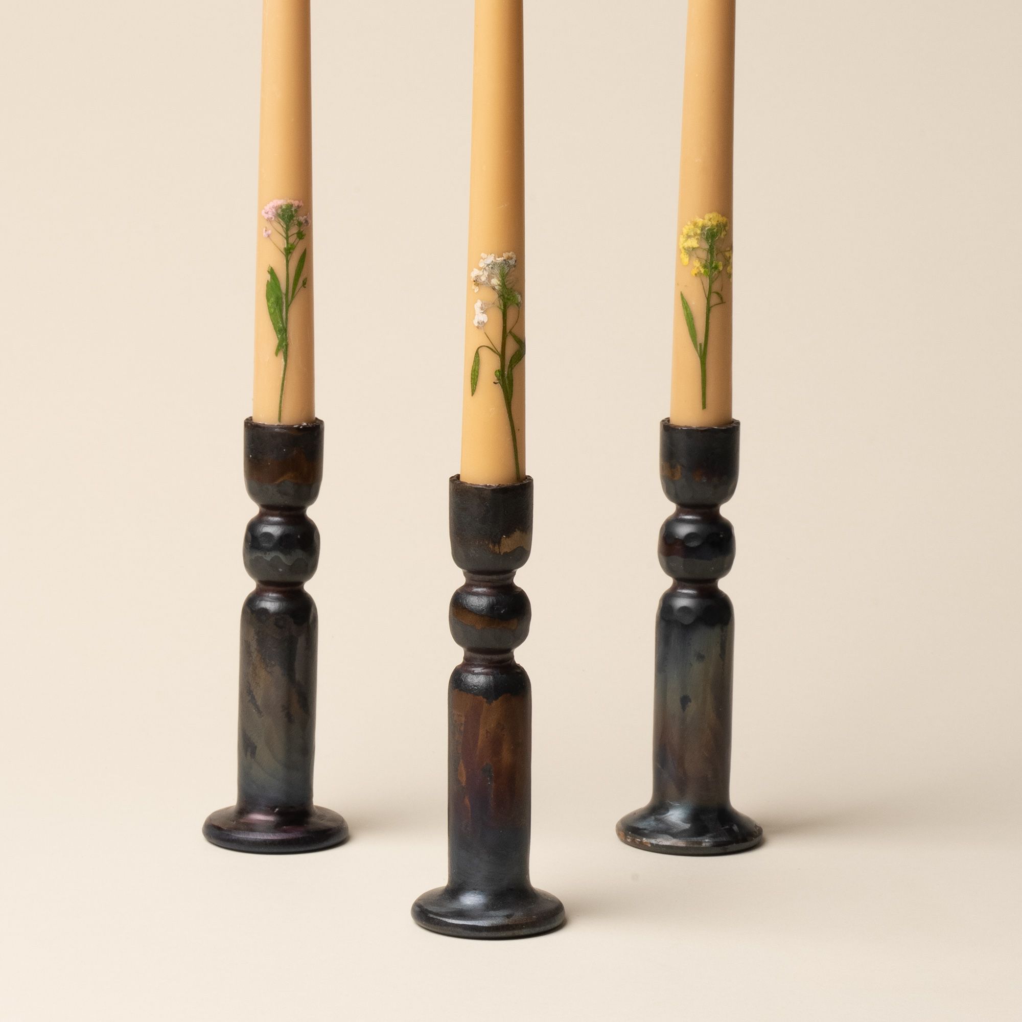 A close up of 3 taper candles with pressed flowers on the bottom, all 3 sitting in a brutalist-styled iron candleholder.