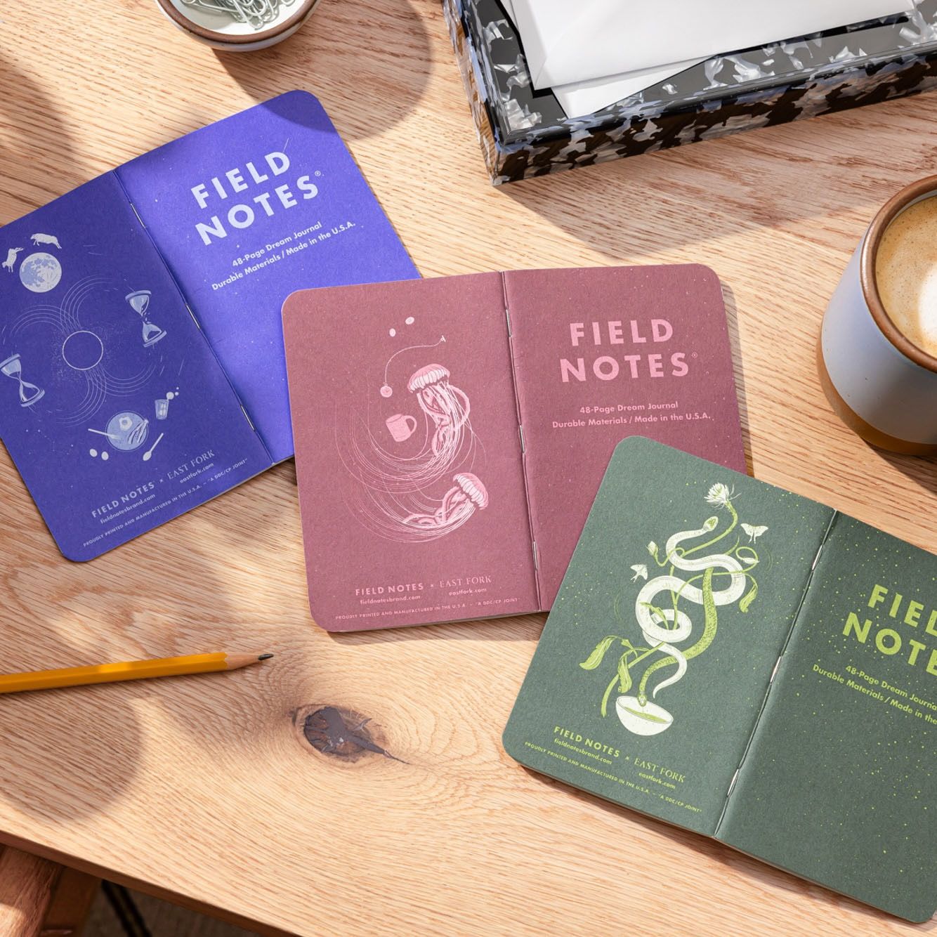 A 3 pack of Field Notes Journal are folded facing down on a desk featuring 3 illustrated back covers in a lapis blue, a plum, and an earthy green.