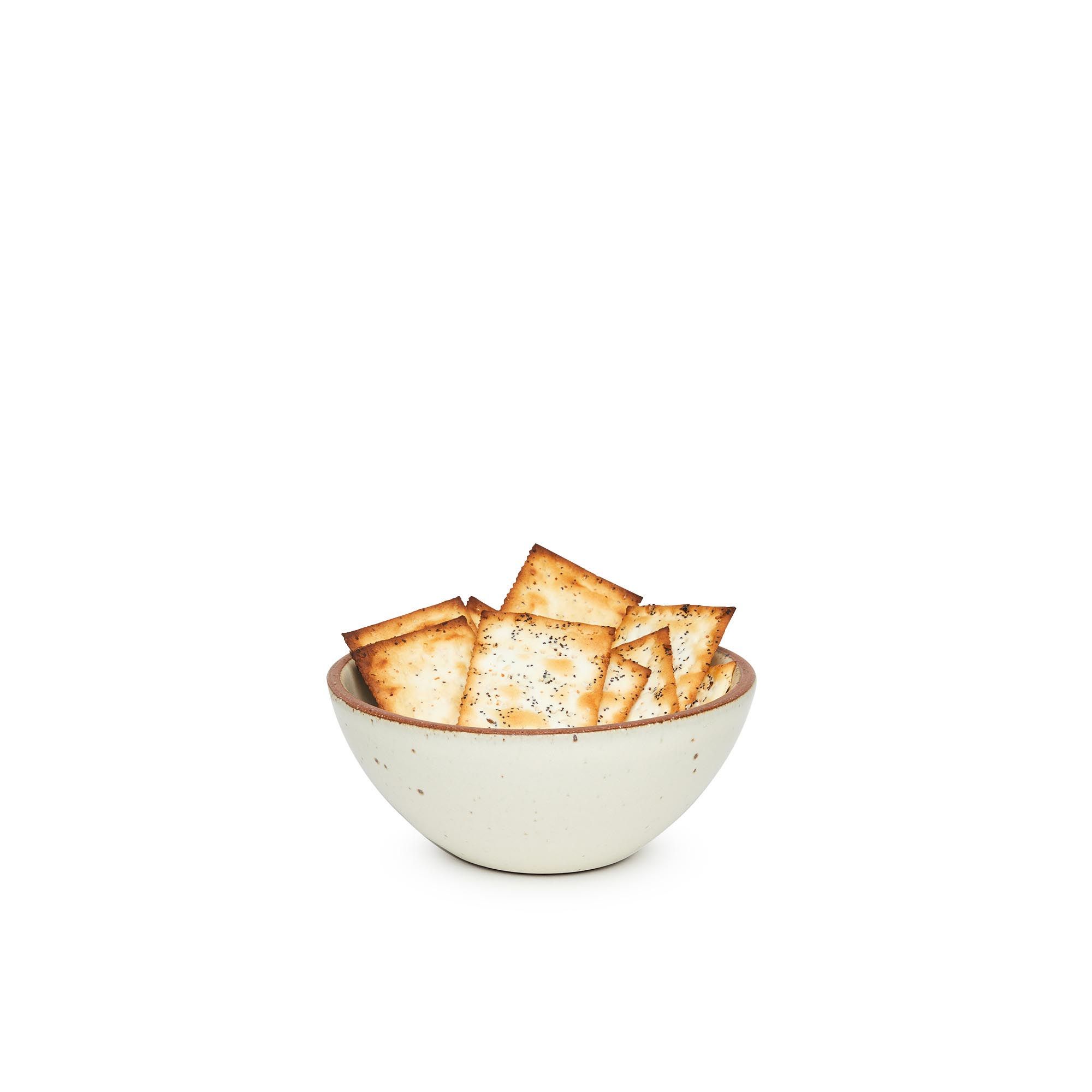 A small dessert sized rounded ceramic bowl in a warm, tan-toned, off-white color featuring iron speckles and an unglazed rim, filled with crackers