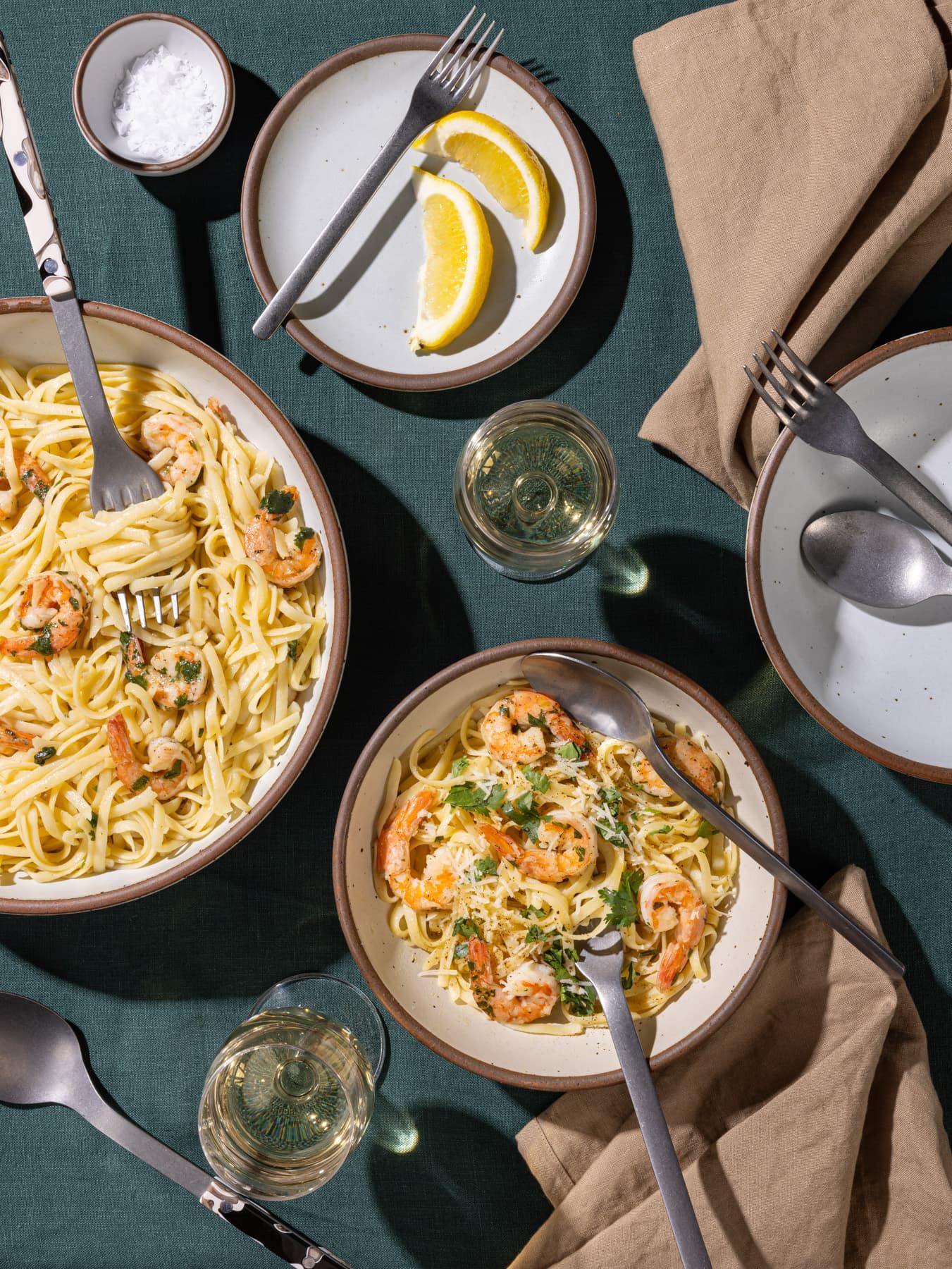 Ceramic bowls in neutral colors with shrimp scampi pasta, steel flatware, lemon wedges, drinks, and dinner napkins.