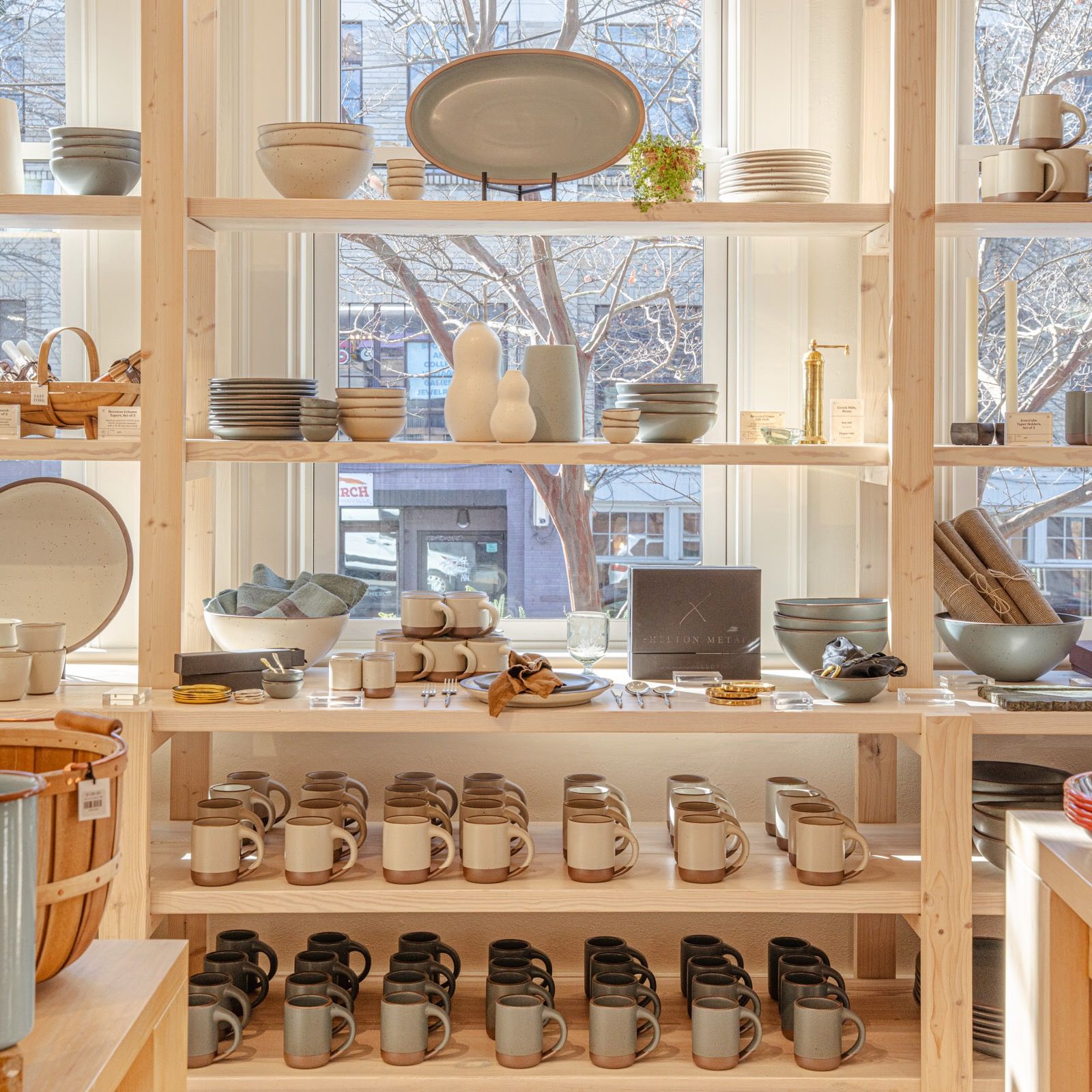 A large wooden shelf piece against a window filled with cream and blue-grey ceramic dinnerware and accessories for the table and kitchen.