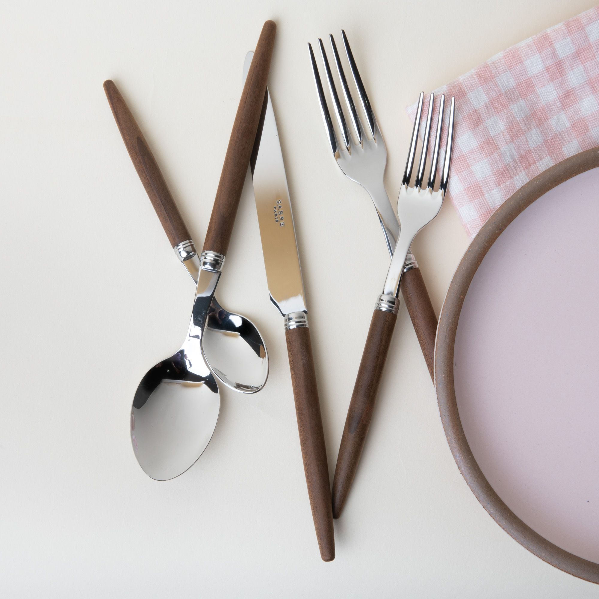 A flatware set of a salad fork, dinner fork, spoon, teaspoon, and knife - all with steel top and simple wood handles. Sitting next to a baby pink plate and a pink and white gingham napkin.