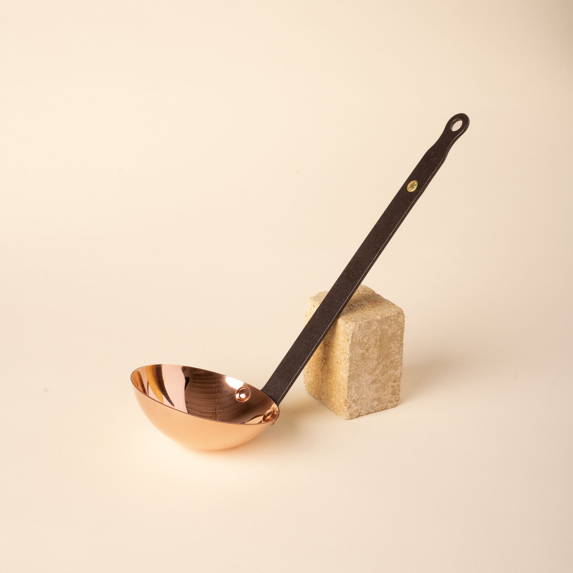 Copper ladle with a long black handle, resting against a textured beige stone block on a cream background.