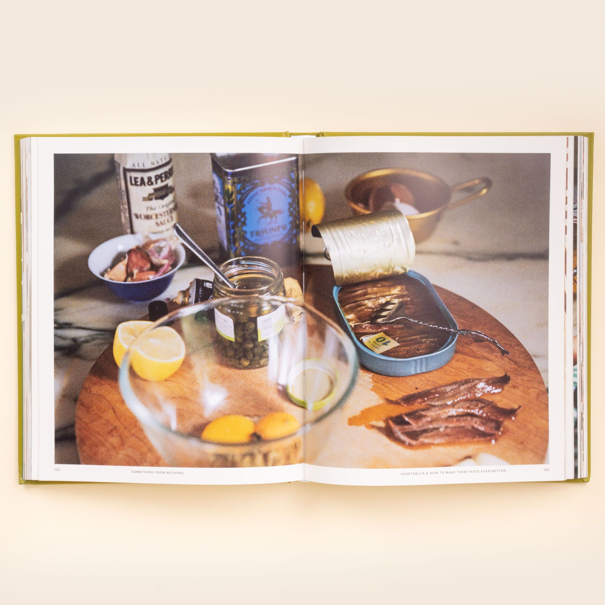 Open cookbook spread showing anchovies, lemons, capers, and seasonings arranged on a wooden cutting board for a recipe prep scene.