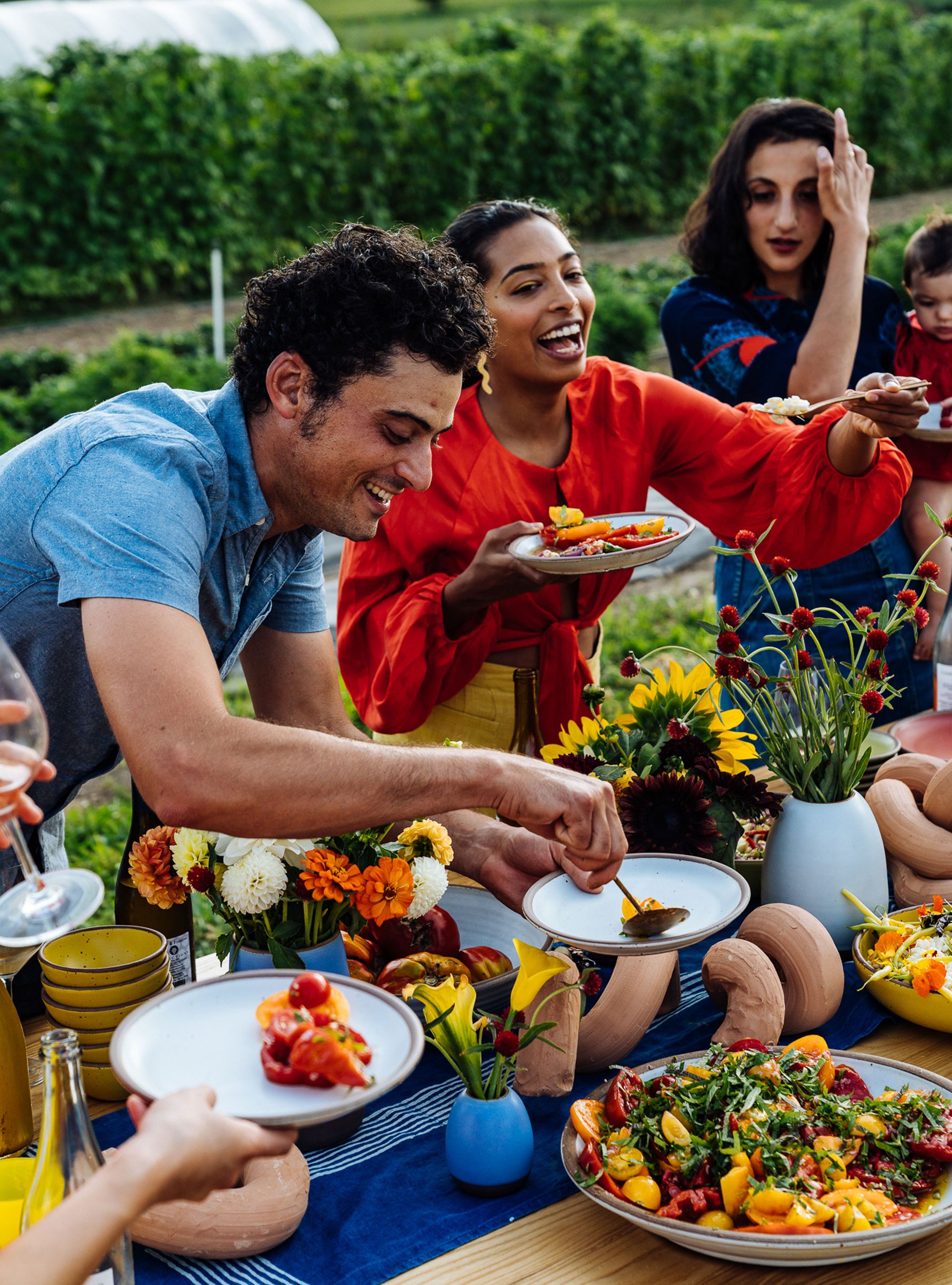 People are smiling outside surrounding a table and filling their plates with beautiful colorful foods.