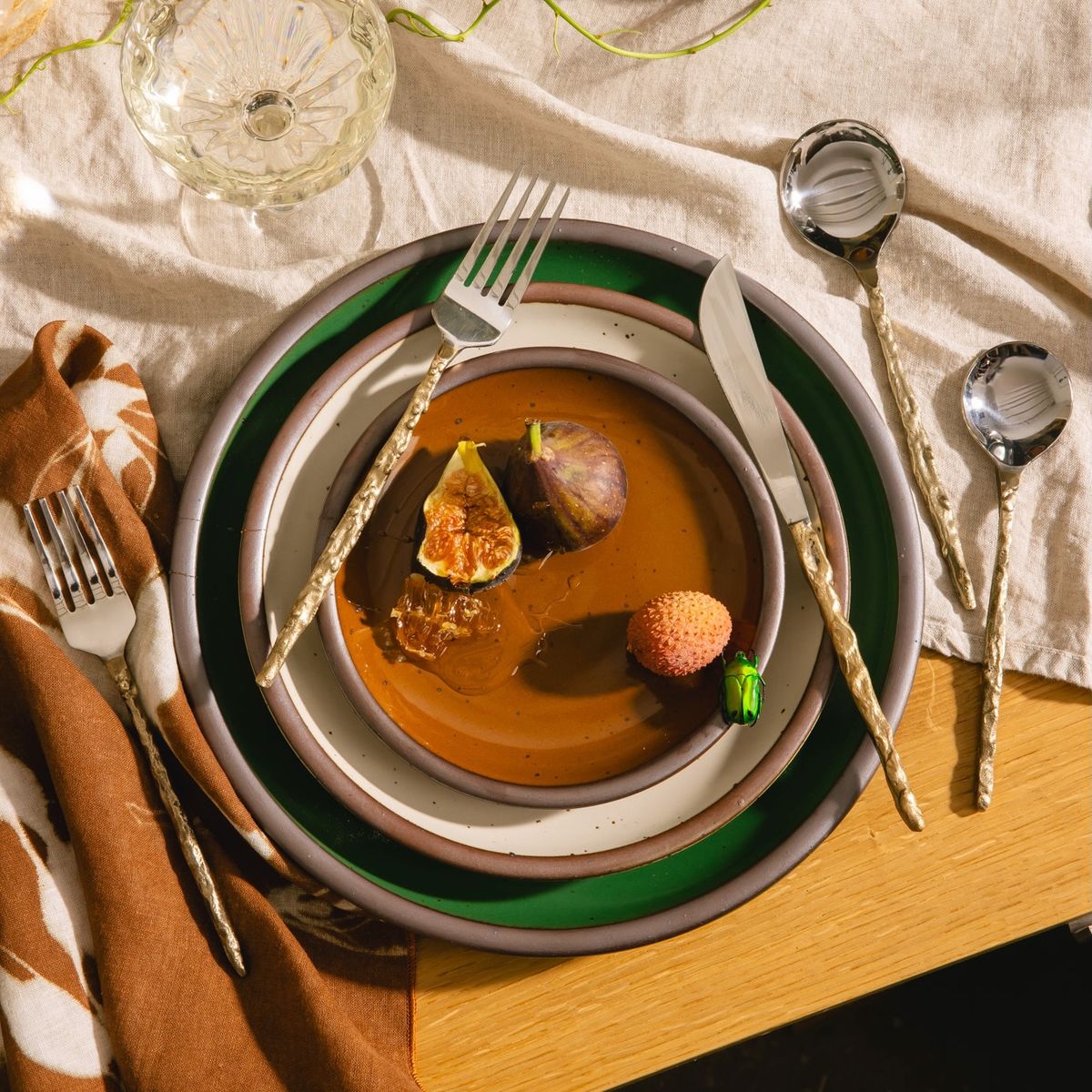 Place setting with gold flatware, stacked plates, figs, lychee, and a green beetle on a linen-draped wooden table.