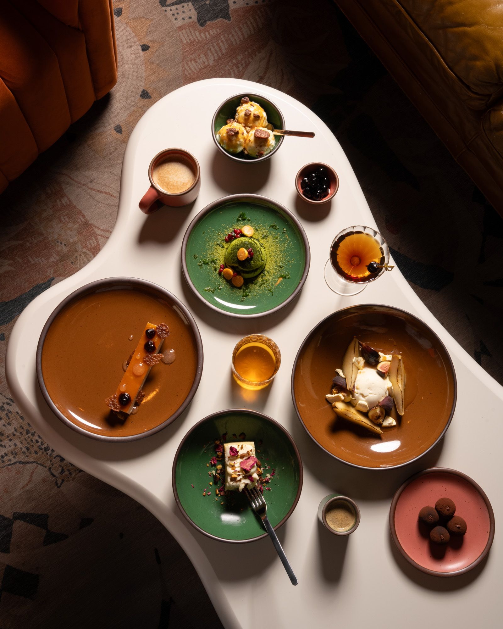 Assorted plated desserts, drinks, and coffee arranged on an organic-shaped white table, featuring glossy green, glossy brown, and pink ceramic dinnerware.