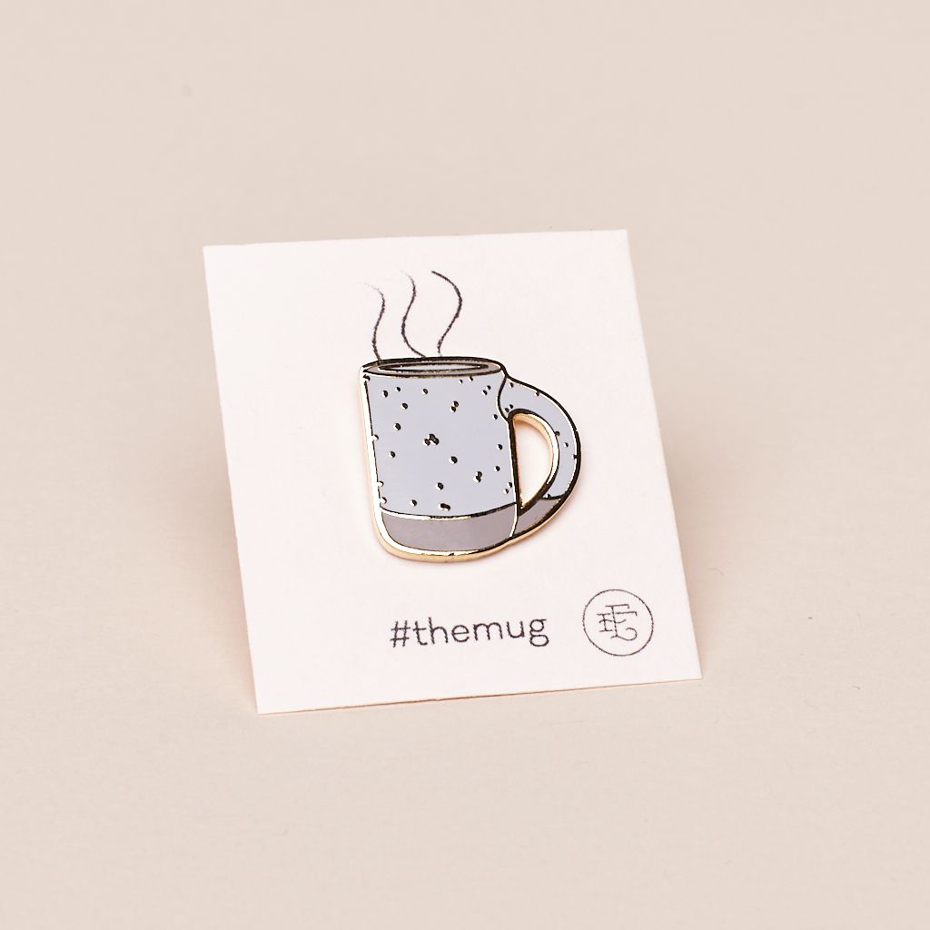 Small enamel pin of a mug illustration with gold outline, gold speckles throughout, and pale pink base