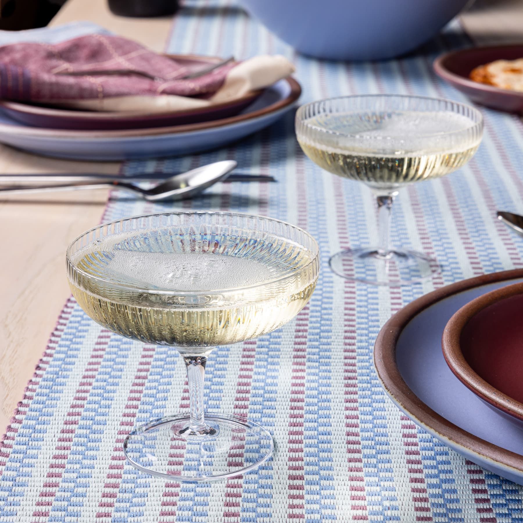 A ribbed traditional coupe glass filled with champagne sitting on a striped periwinkle table runner.