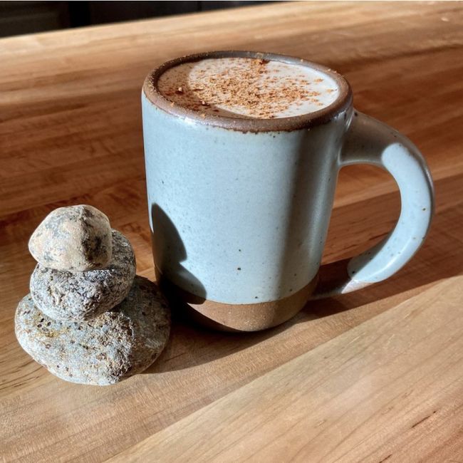 The East Fork Mug in Soapstone sits on top of a wooden tabletop next to a stack of stones from a riverbed.