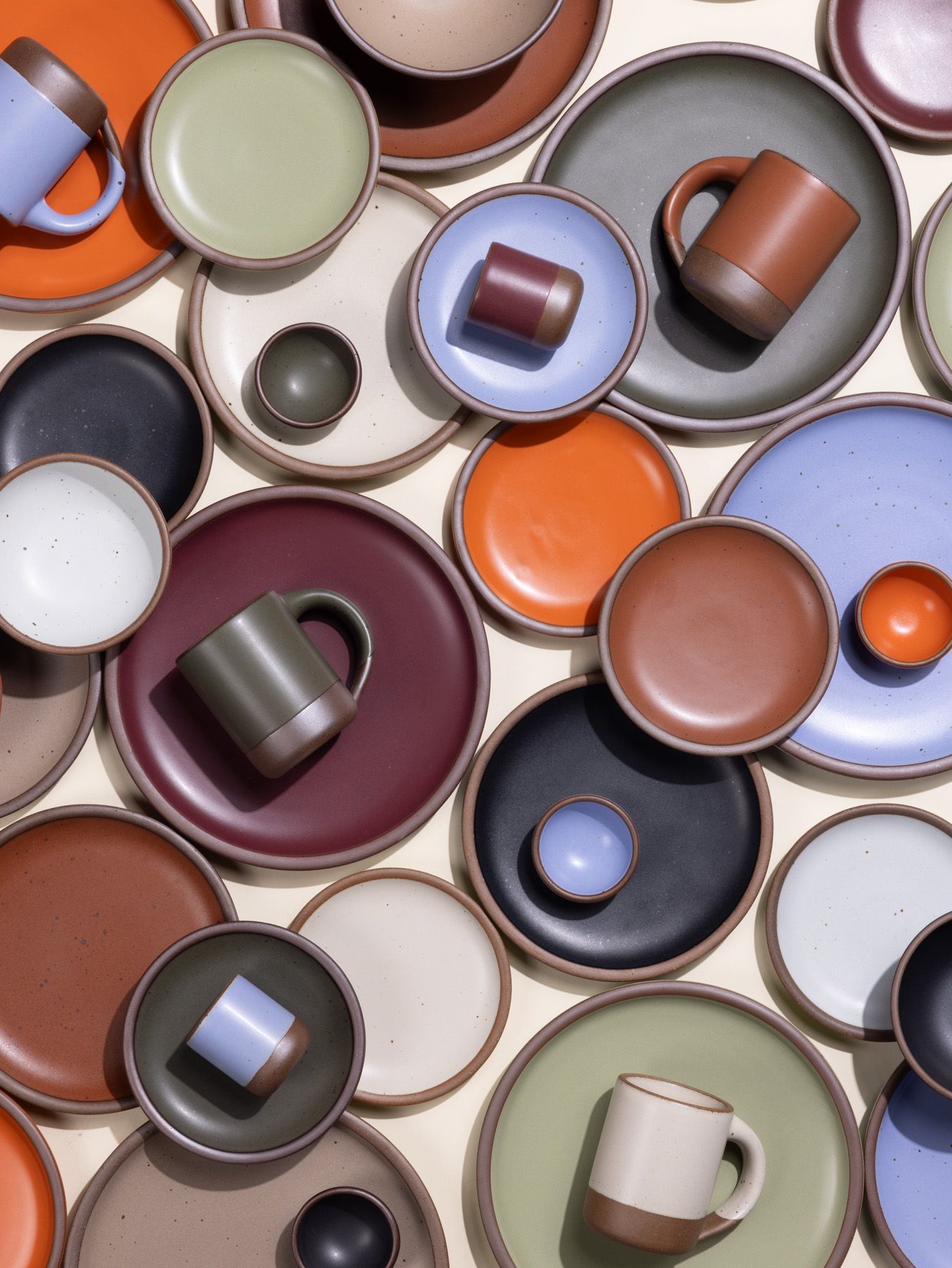 An artful layout of ceramic plates, bowls, and mugs of all different sizes in a variety of colors including plum, cream, black, light sage green, terracotta, bright orange and more.