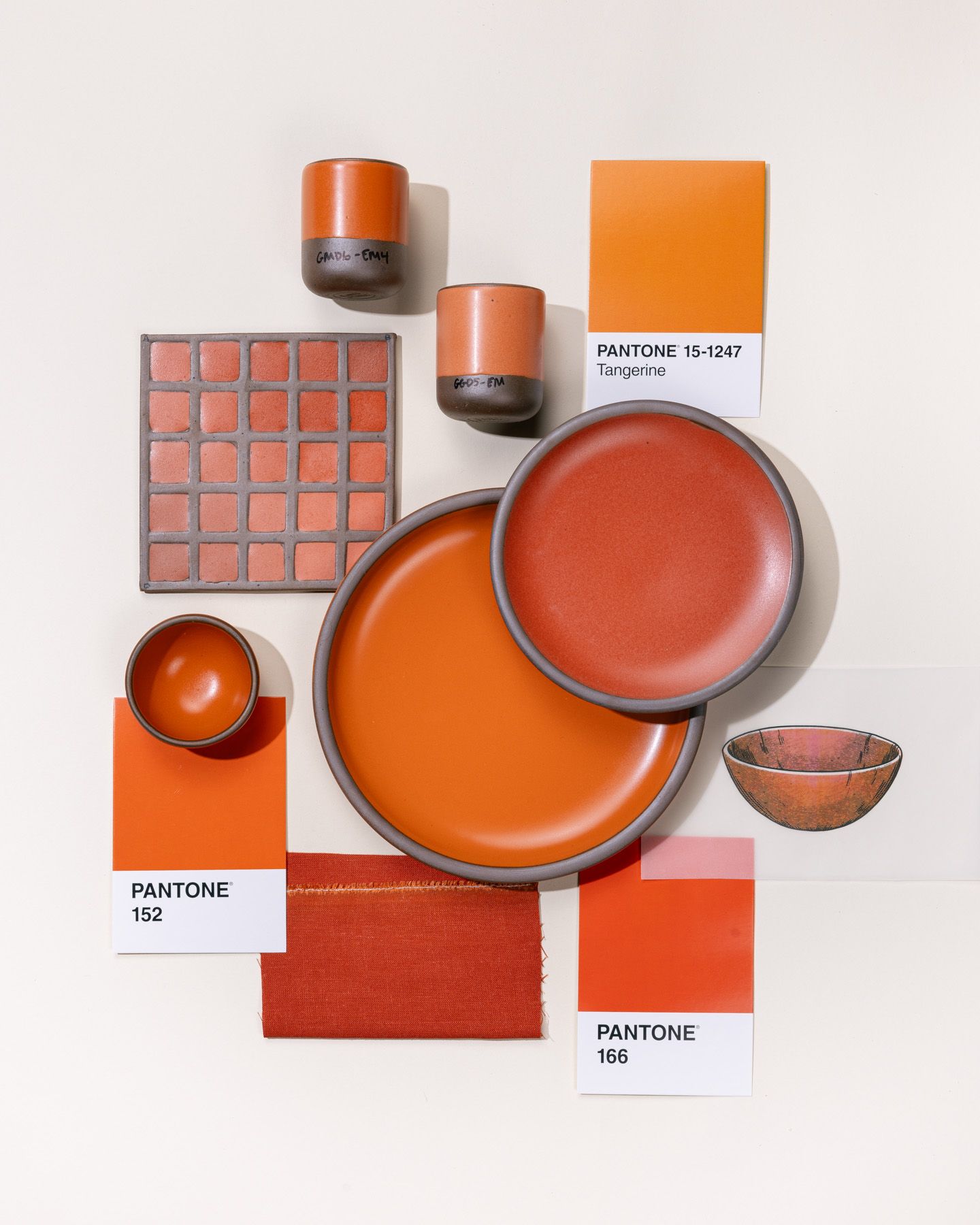 An overhead view of a flat lay featuring 2 ceramic plates in varying bold orange colors, surrounded by pantone color cards, illustrations, and a ceramic color tile also all in orange tones.