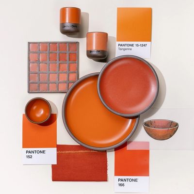 An overhead view of a flat lay featuring 2 ceramic plates in varying bold orange colors, surrounded by pantone color cards, illustrations, and a ceramic color tile also all in orange tones.