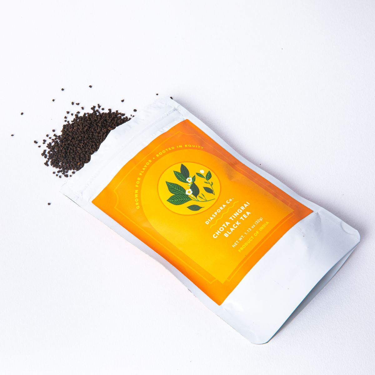 Ground black tea leaves spilled out of an open packaged white bag with an orange label with floral illustration and text that reads "Chota Tingrai Black Tea"