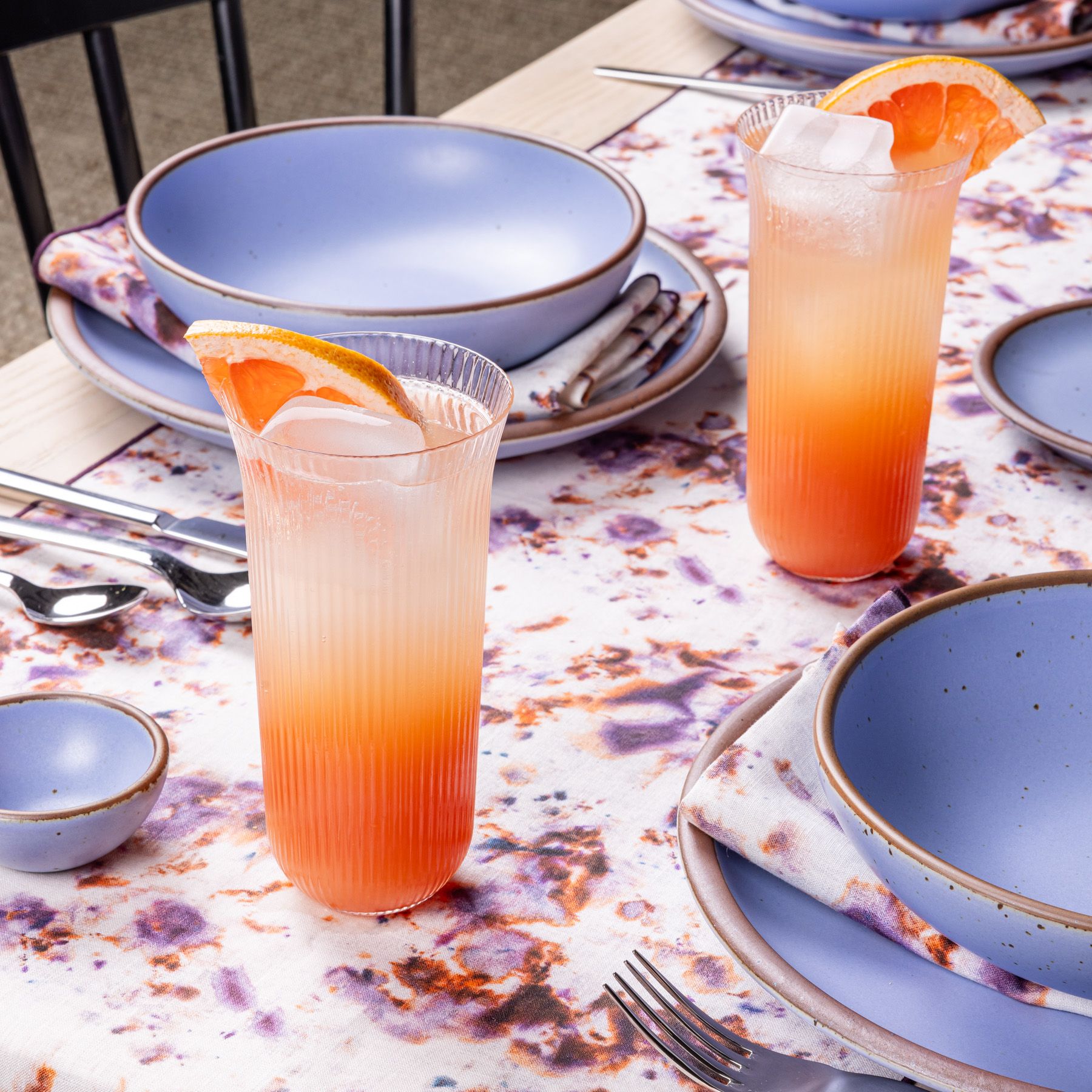 Two peachy, citrus iced drinks on a purple, orange and white marbled tablecloth, along with light periwinkle ceramic place settings at a table. Napkins are laid on the plate matching the marbled tablecloth. 