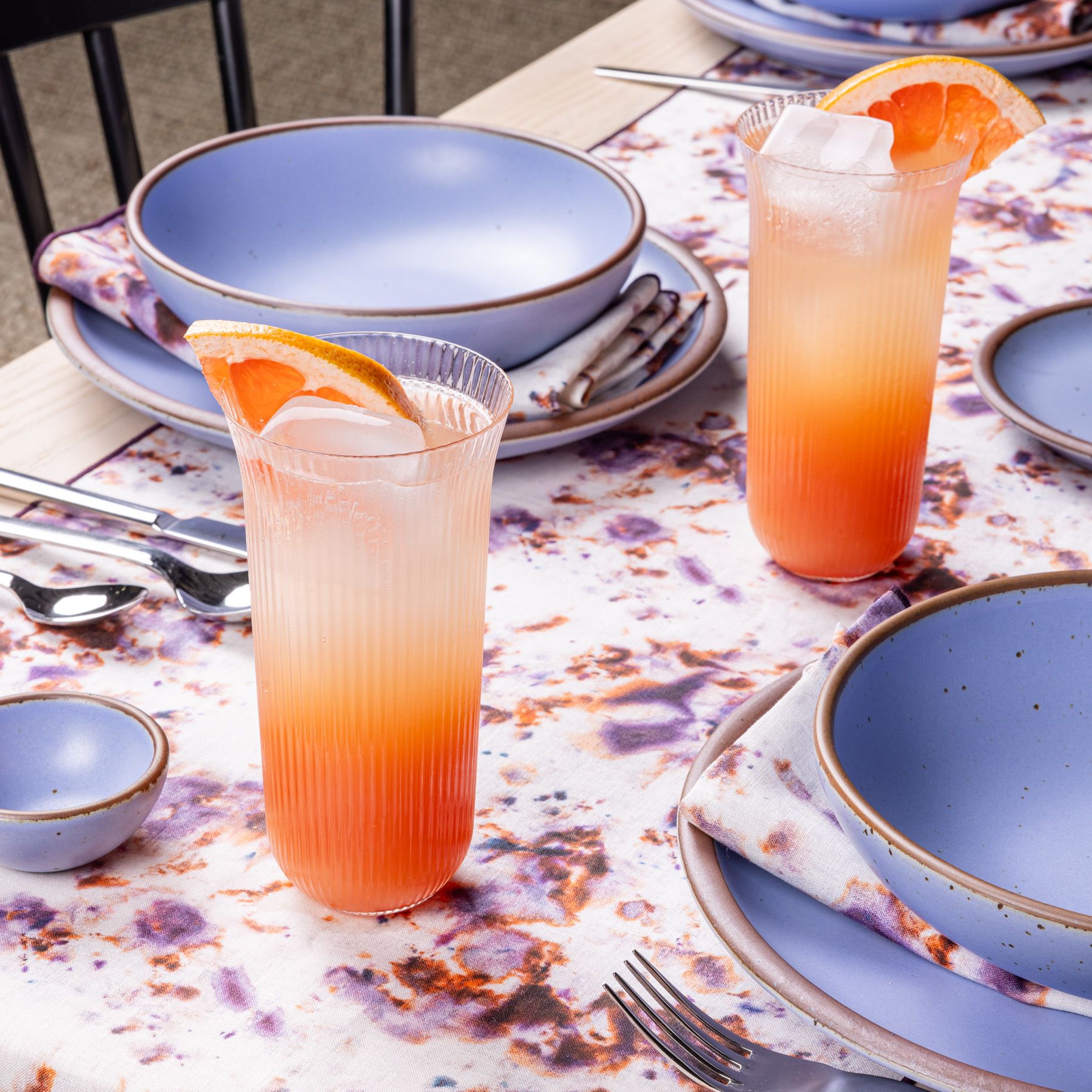 Two peachy, citrus iced drinks on a purple, orange and white marbled tablecloth, along with light periwinkle ceramic place settings at a table. Napkins are laid on the plate matching the marbled tablecloth.