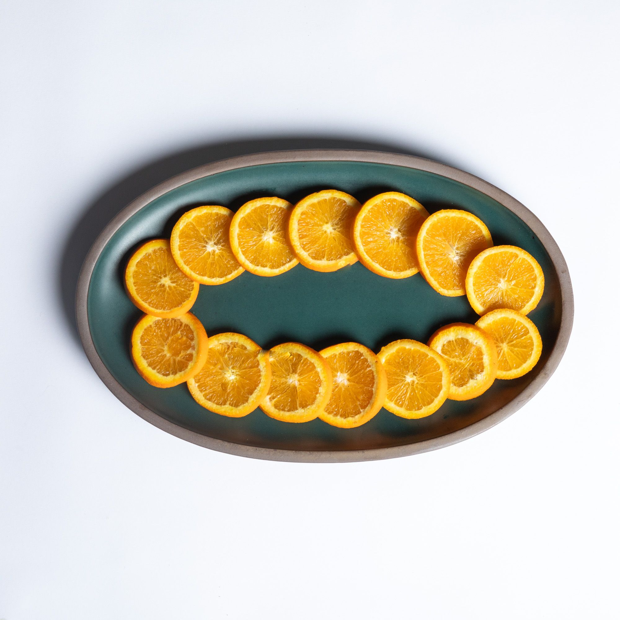 A large oval ceramic platter in a deep, dark teal color featuring iron speckles and an unglazed rim, with an oval of orange slices