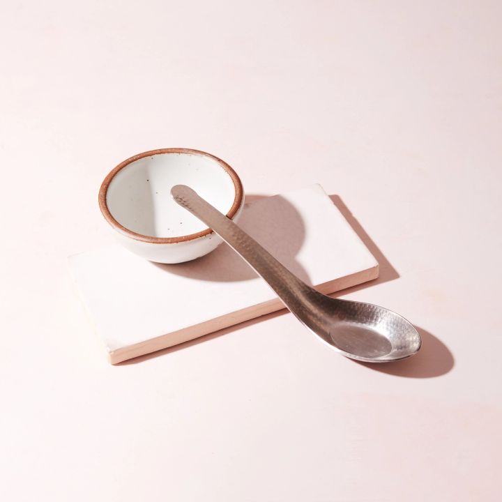 A steel Asian soup spoon with hammered finish leans on a small ceramic white bowl and tray