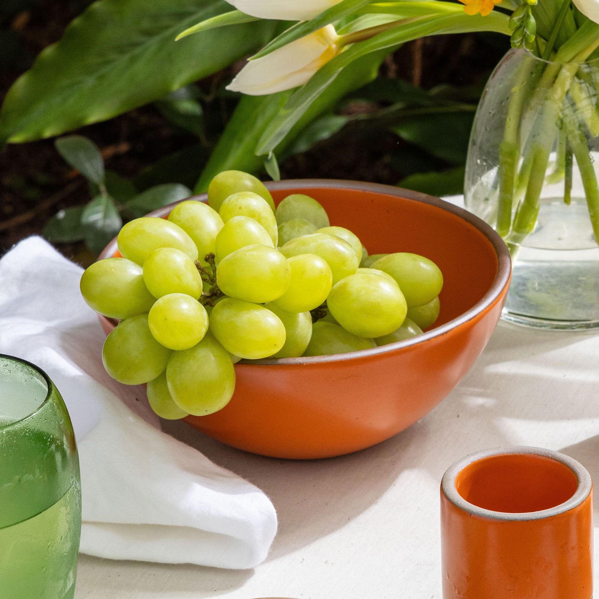 On a table sits a bold orange bowl with green grapes, surrounded by a clear vase, tiny cup, and white napkin