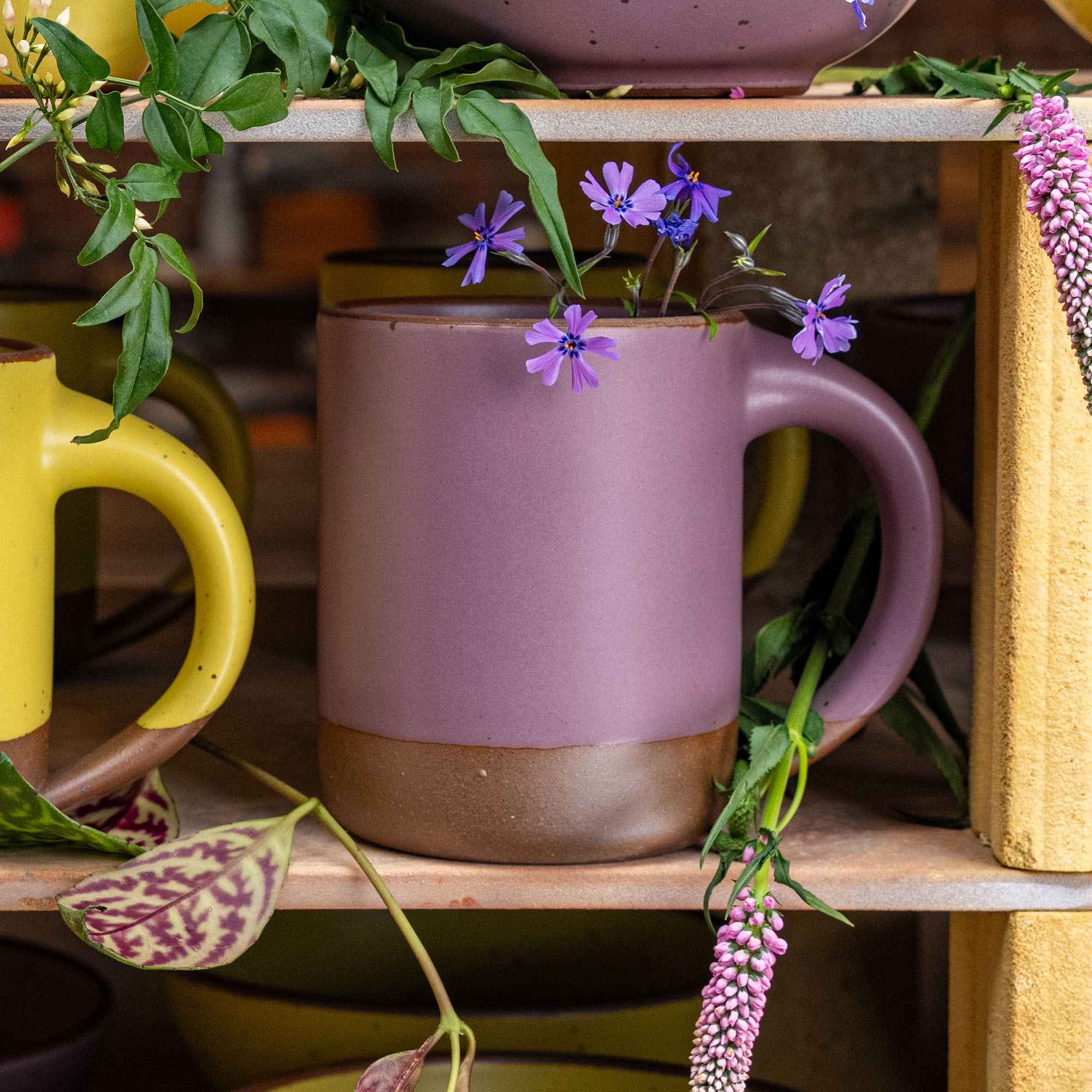 A large sized ceramic mug with handle in a soft mauve purple color featuring iron speckles and unglazed rim, sits on a wood shelf and is surrounded by flowers