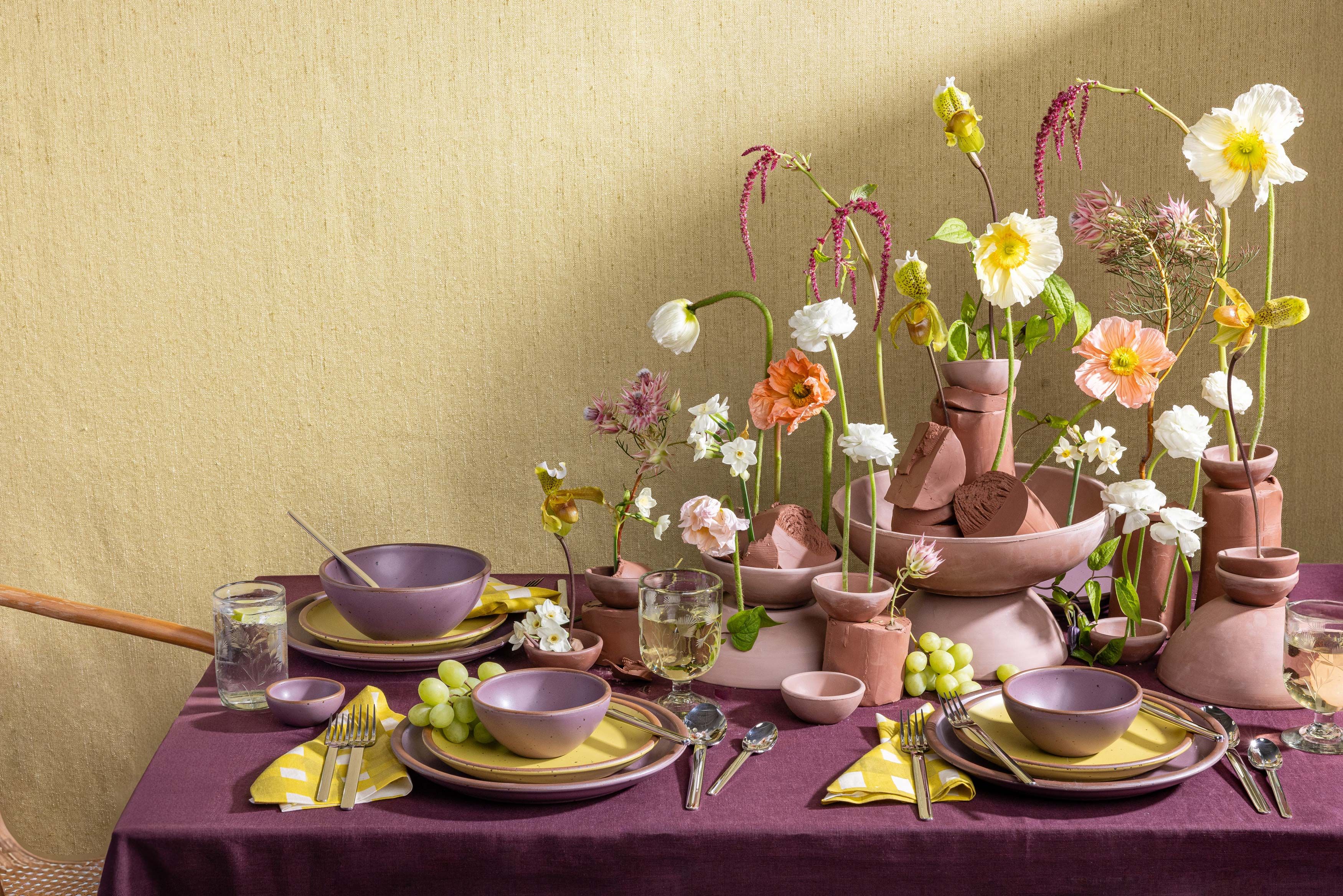 A beautiful tablescape filled with zesty yellow and soft mauve purple ceramic dinnerware, purple tablecloth, and large and lively floral sculpture centerpiece