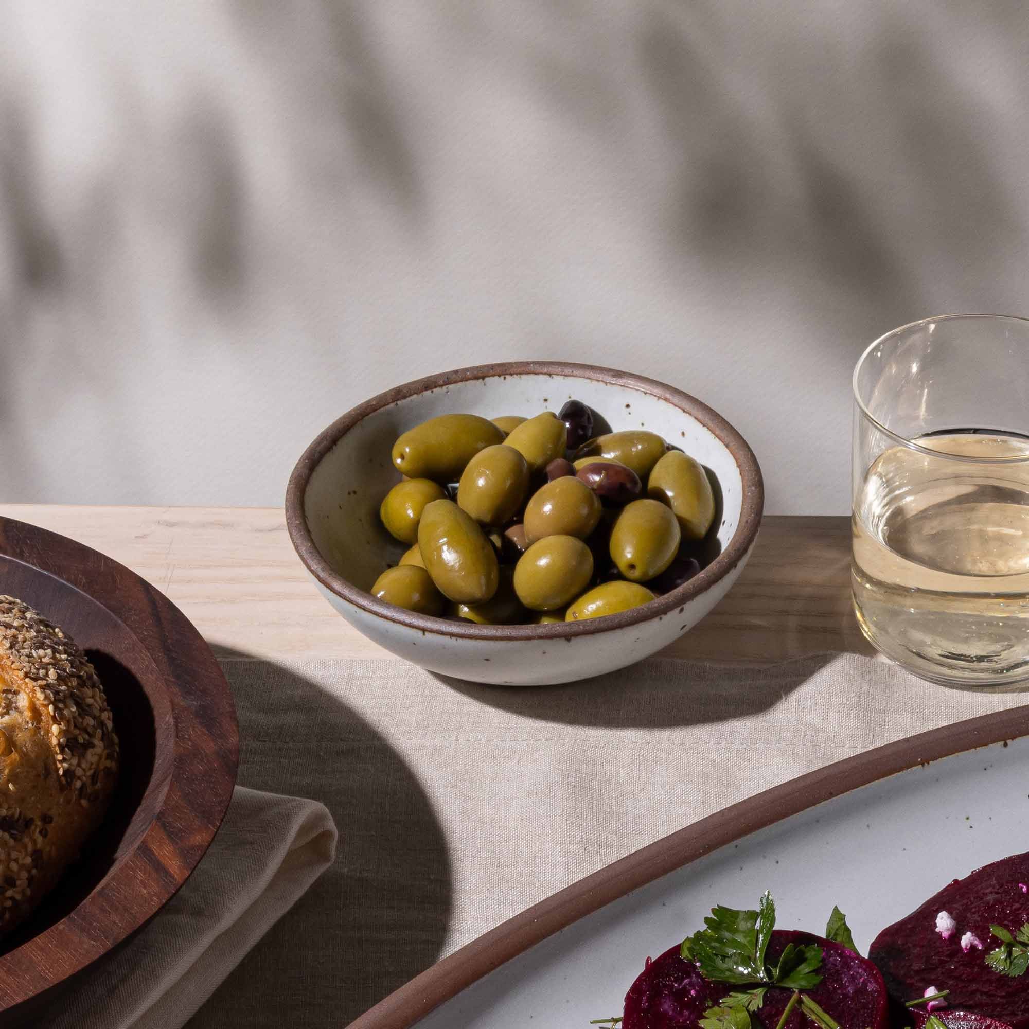 A shallow ceramic bowl in a cool white color with olives inside and more plates, glassware, and a wooden bowl nearby.