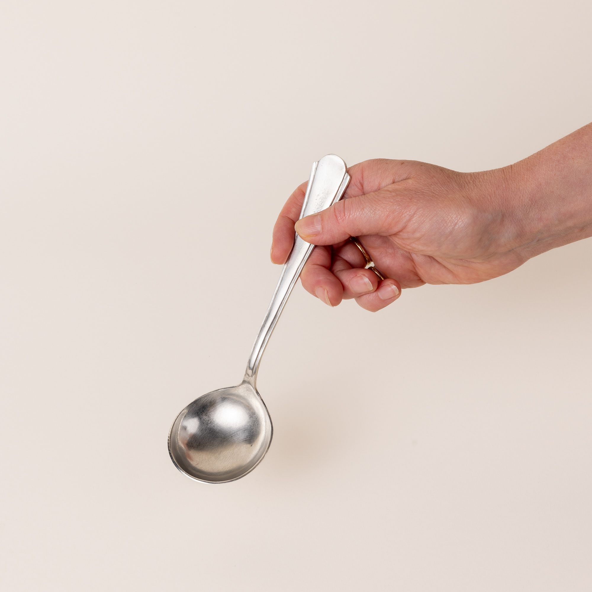 A hand holding the handle of a silver gravy spoon, with a skinny handle that gets a little wider and round on the end. The bowl of a spoon is circular.