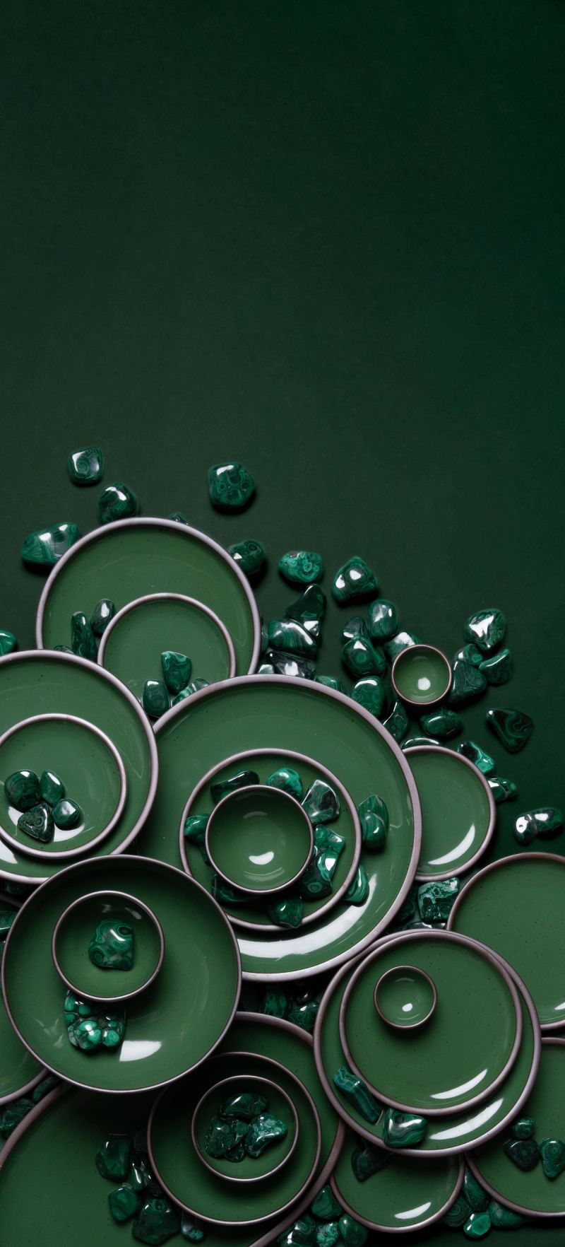 Arrangement of glossy emerald green ceramic plates and bowls with scattered malachite stones on a dark green background.