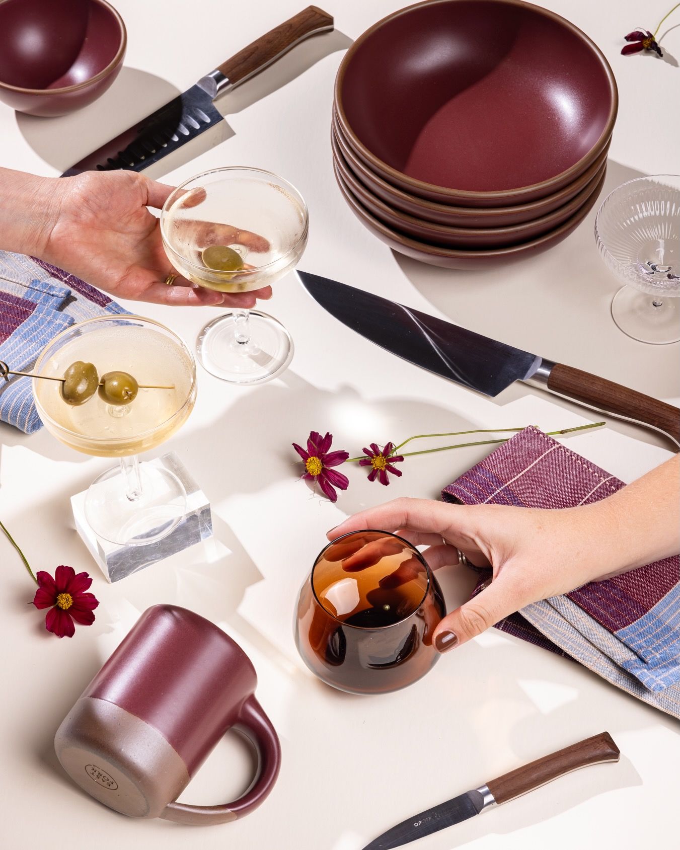 Hands reach for a martini coupe and a whiskey snifter against a white background. All around are other items including a stack of shallow bowls, kitchen knives, a plum ceramic mug, and more.