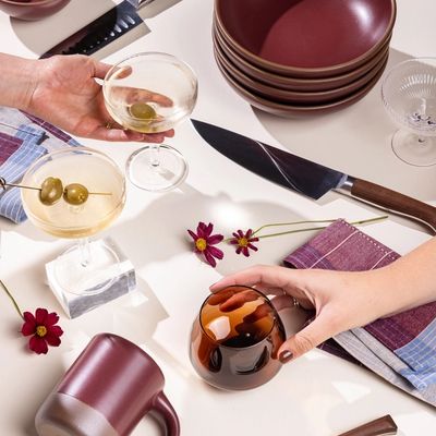 Hands reach for a martini coupe and a whiskey snifter against a white background. All around are other items including a stack of shallow bowls, kitchen knives, a plum ceramic mug, and more.