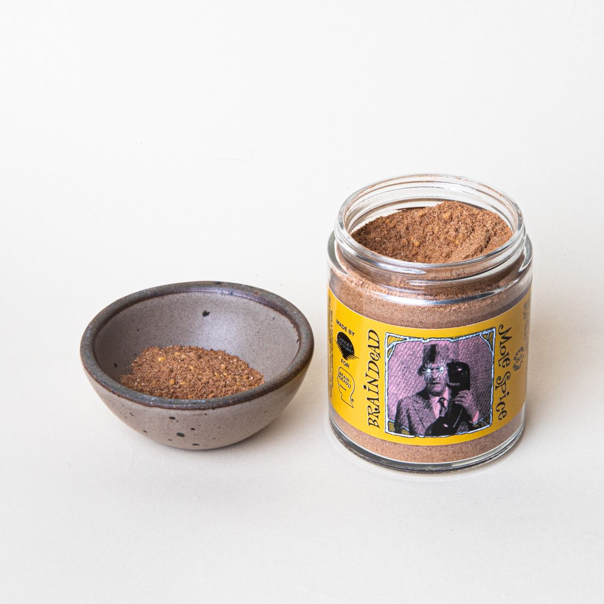 An open short cylinder jar with a butter yellow label that reads "Braindead Mole Spice" and a tiny bowl with spice inside next to it