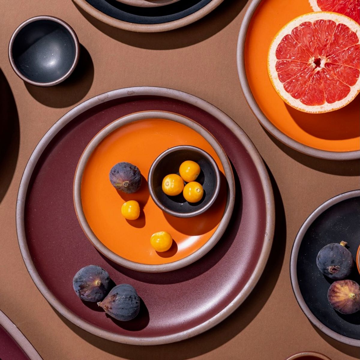 An artful arrangement of a bold orange side plate, a plum dinner plate, and tiny black bowls with colorful fruits