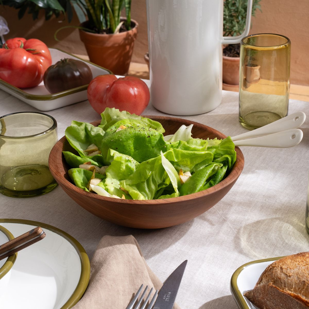 Fresh green salad in a wooden bowl on a table set with enamelware, glassware, and heirloom tomatoes in natural light.