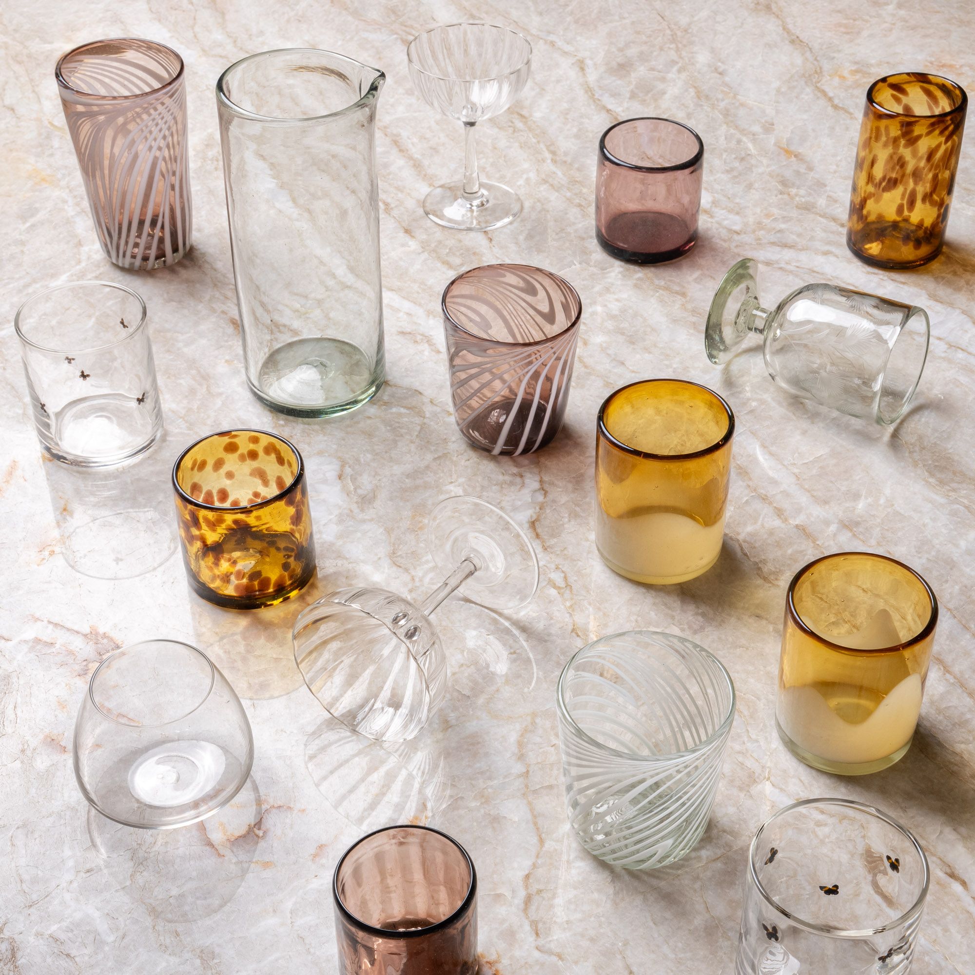 Assorted handblown drinking glasses and stemware in clear, amber, and patterned glass arranged on a marble surface.
