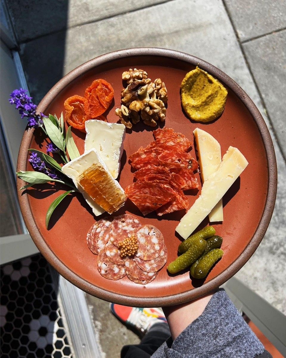 A hand holds a terracotta ceramic plate filled with charcuterie