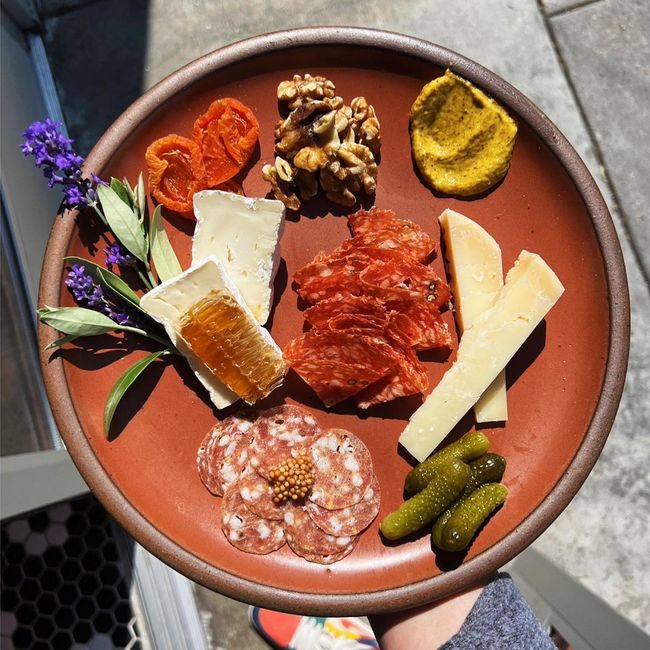 A hand holds a terracotta ceramic plate filled with charcuterie