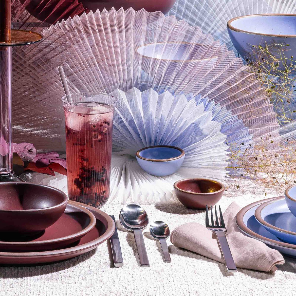 A delicate ribbed tall glass that curves out on the top with ceramic plum dinnerware, artful arrangement, and silverware.