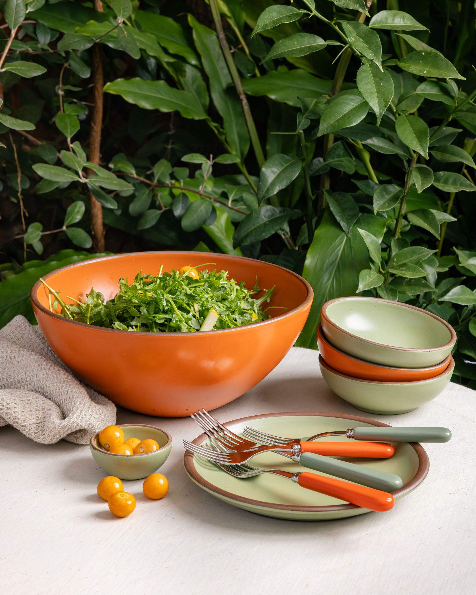 In an outdoor setting, a large bowl, smaller bowls, and side plate in bold orange and sage green colors sits with flatware of the same color palette.