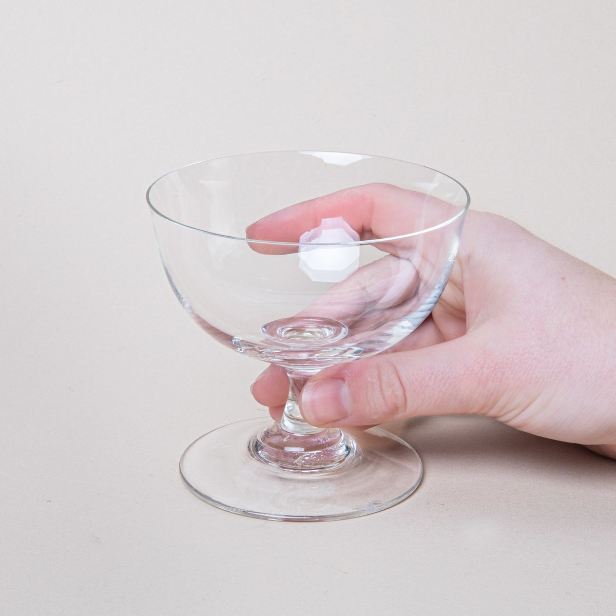 A hand holding a clear coupe glass with a short stem and wide base