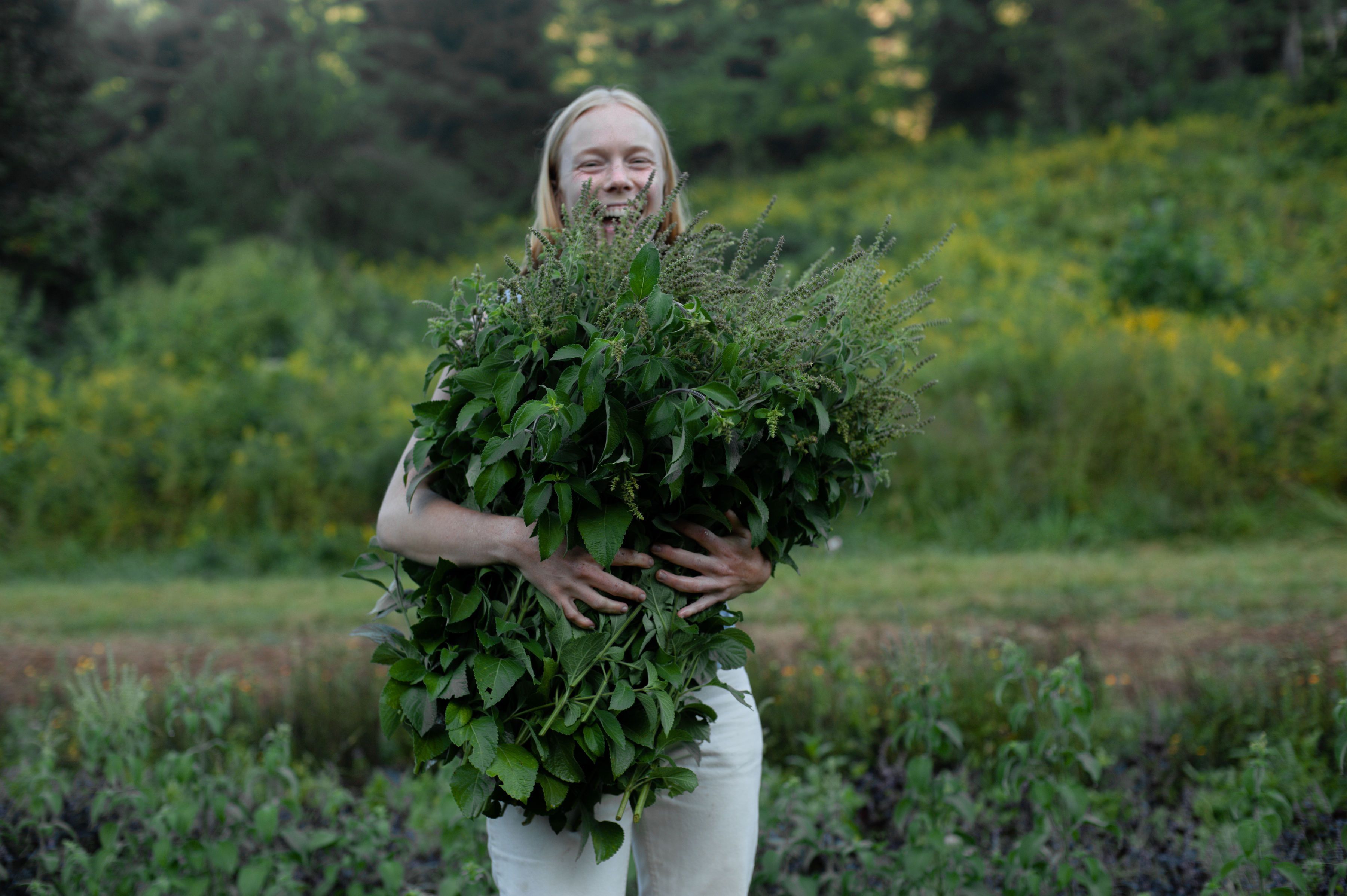 Outside, a woman holds a large bushel of greenery and herbs and is smiling at the camera.