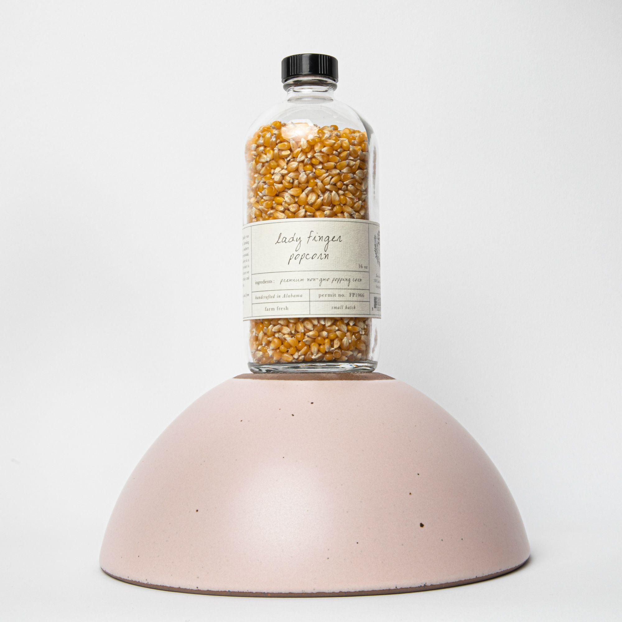 A glass bottle of popcorn kernels with an off white label that reads "Lady finger popcorn" sits on top of an upside down popcorn bowl.